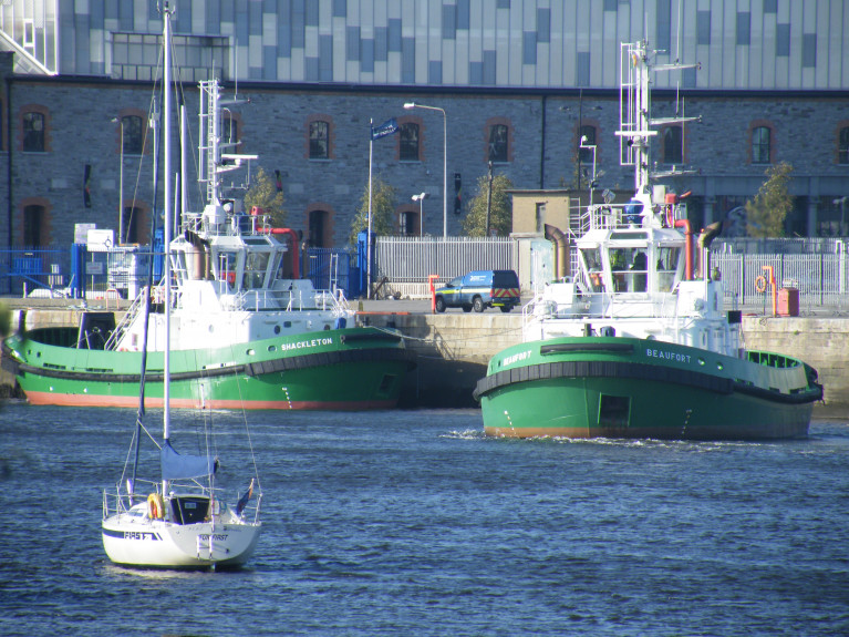 Towage Twins Relocate: Dublin Port owned tugs, Shackleton berthed on left while Beaufort sets off on towage duties from their previous station berth located on the North Quay Wall Extension next to the Tom Clarke Bridge (East-Link) not in shot but the Three Arena is seen above. Now the tugs have a more centrally located station berth downriver to faciliate easier operations and for the safety of crew using a custom built pontoon. Note, the tugs don&#039;t feature funnel&#039;s, instead use exhaust (pipes) uptakes positioned on both sides of the tug master&#039;s wheelhouse. 